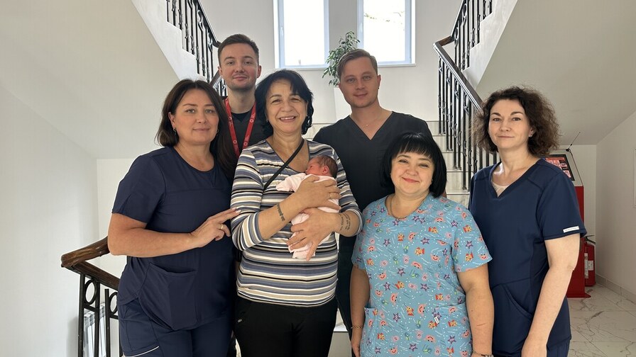 У лікарняному холі Наталя тримає немовля на руках, біля неї стоять медичні працівники, які супроводжували вагітність та приймали пологи